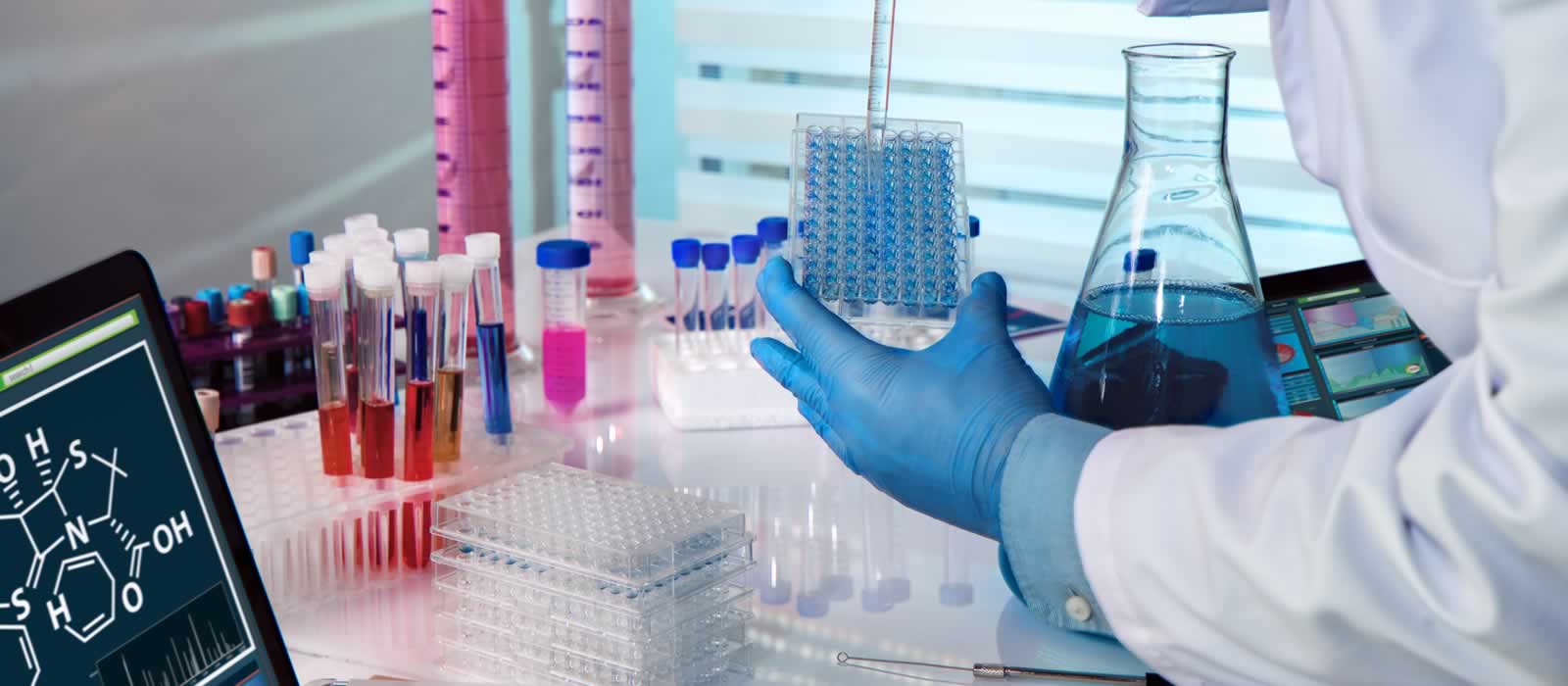 A scientist in white coat and blue safety gloves fills a samples tray with dyed liquid prior to studying it under a microscope.