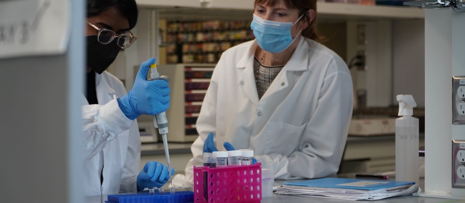 A white, female student watches a man of colour - a professor or a lab technician - fill test tubes with samples. 
